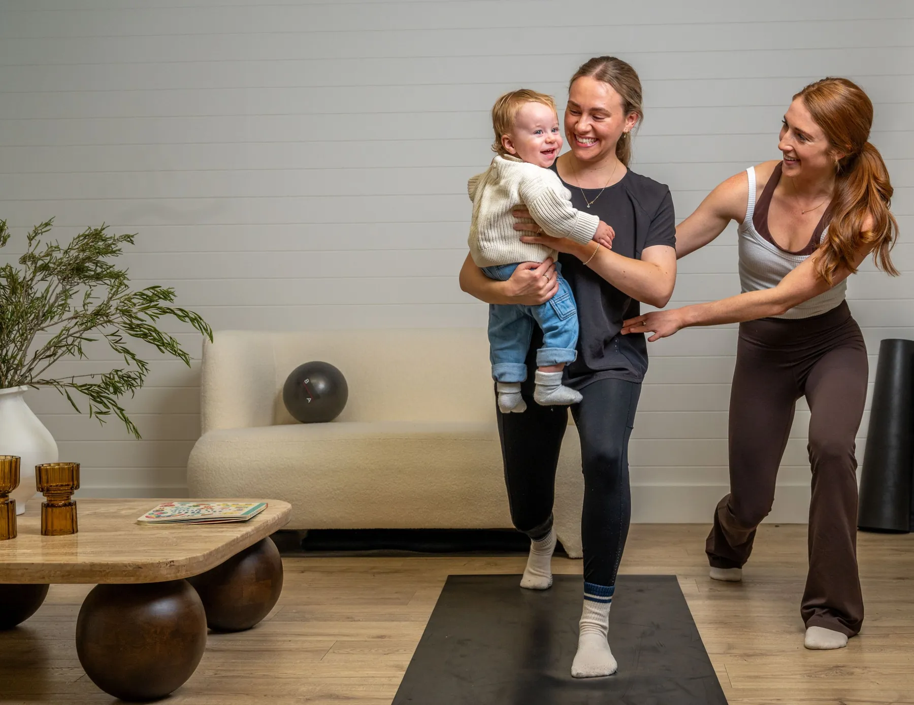 Bridget guiding a new mum through a lunge while she holds her baby at home